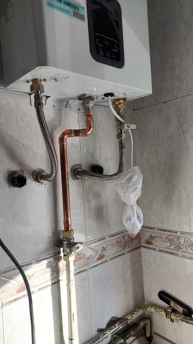 Calentador blanco recién conectado sobre azulejo claro, con la tubería de cobre, los latiguillos y la llave de gas debajo.