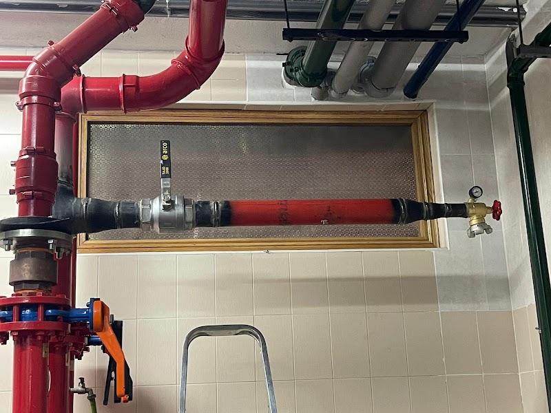 Tubería de gas pintada de rojo en una sala de calderas, con una válvula, un manómetro y una escalera de mano debajo.