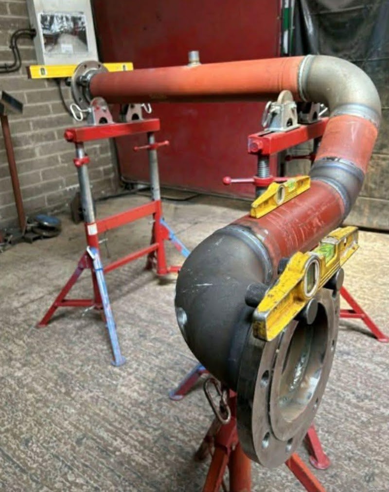 Tramo de tubería de acero con un codo y una brida, apoyado en caballetes rojos con dos niveles encima, en un taller.
